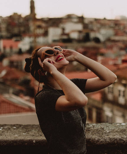 Image of a woman wearing sunglasses and looking up to the sky in a city in Europe - KILROY