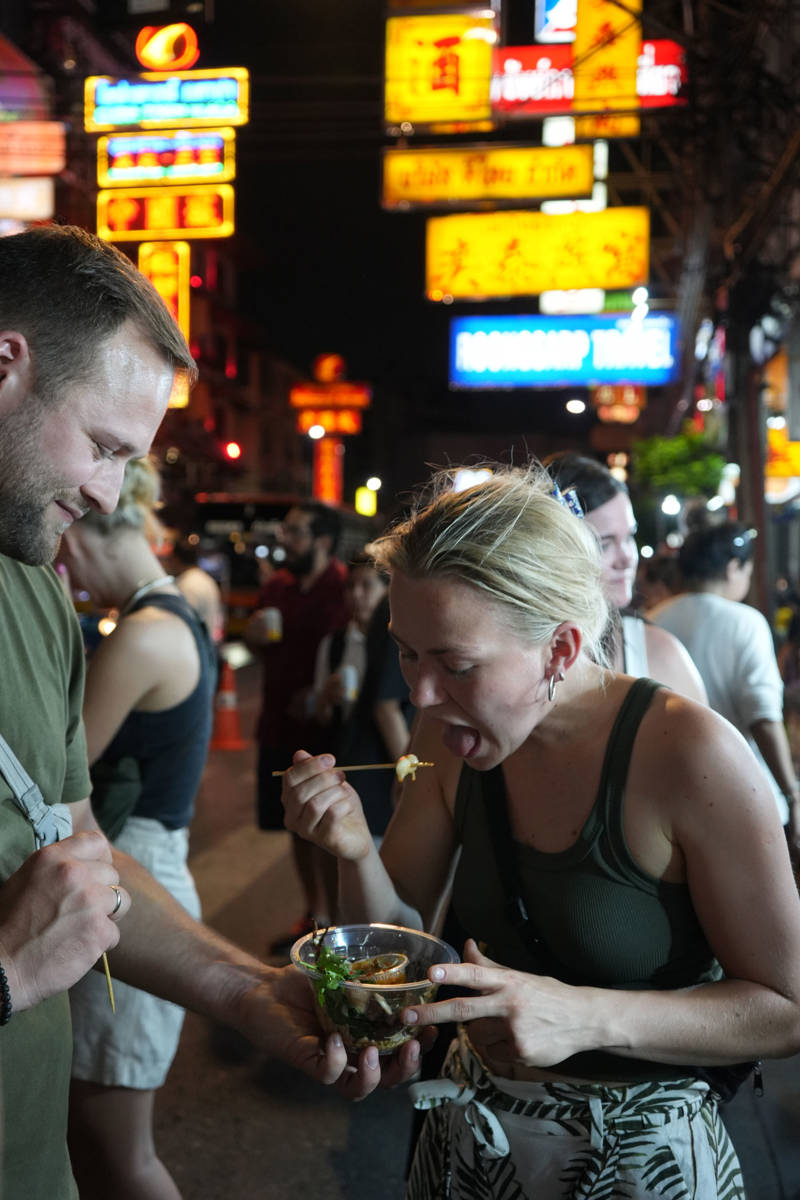 Group of KILROY travellers exploring a night market in Bangkok and trying streetfood, eating with sticks from a plastic cup